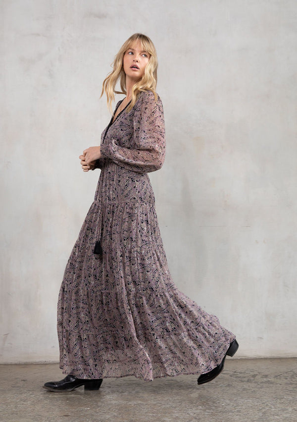 [Color: Black/Orchid] A model wearing a sheer black and orchid paisley print bohemian maxi dress. With contrast crochet trim, a tiered skirt, long voluminous sleeves with smocked elastic wrist cuffs, and an open back with a self covered button closure.