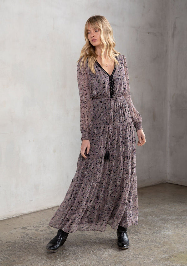 [Color: Black/Orchid] A model wearing a sheer black and orchid paisley print bohemian maxi dress. With contrast crochet trim, a tiered skirt, long voluminous sleeves with smocked elastic wrist cuffs, and an open back with a self covered button closure.