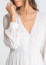 [Color: Gardenia] A model wearing a dreamy sheer off white maxi dress in swiss dot chiffon. With lace trim details throughout, a self covered button front, a sexy front slit, and sheer voluminous long sleeves with a button wrist cuff.