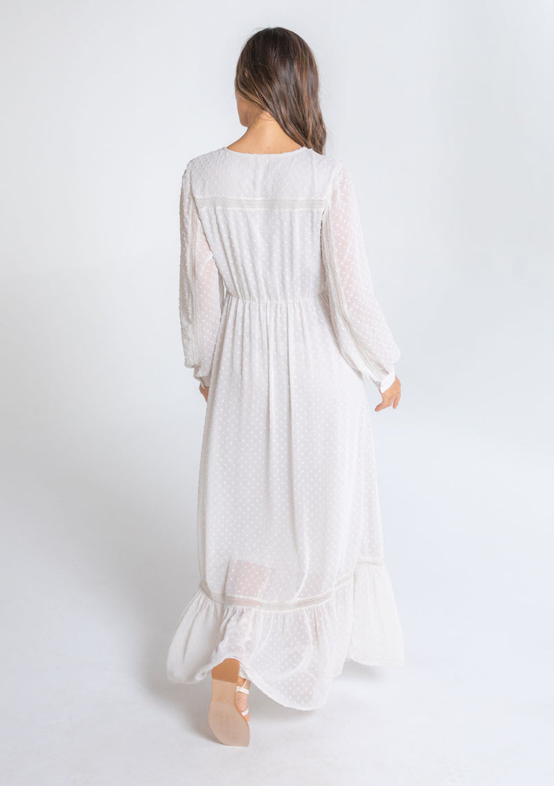 [Color: Gardenia] A model wearing a dreamy sheer off white maxi dress in swiss dot chiffon. With lace trim details throughout, a self covered button front, a sexy front slit, and sheer voluminous long sleeves with a button wrist cuff.