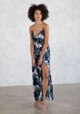 [Color: Navy/Lilac] A model wearing a sexy navy and lilac purple silky cowl neck maxi dress in a large floral print. With a half elastic waist at the back, a side slit, and an adjustable strappy back. 