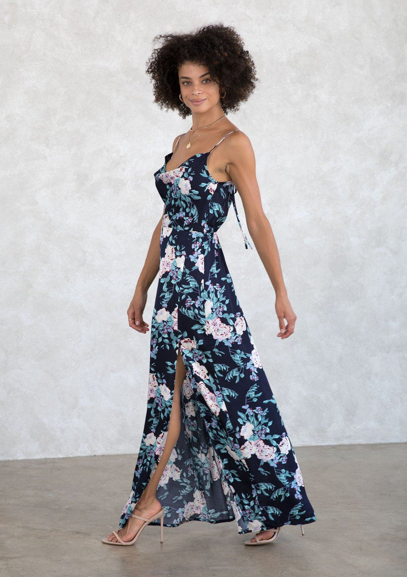 [Color: Navy/Lilac] A model wearing a sexy navy and lilac purple silky cowl neck maxi dress in a large floral print. With a half elastic waist at the back, a side slit, and an adjustable strappy back. 