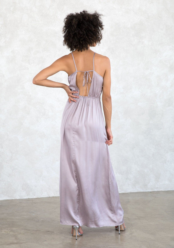 [Color: Lilac Mist] A model wearing a sexy silky lilac purple cowl neck maxi dress. With a half elastic waist at the back, a side slit, and an adjustable strappy back. 