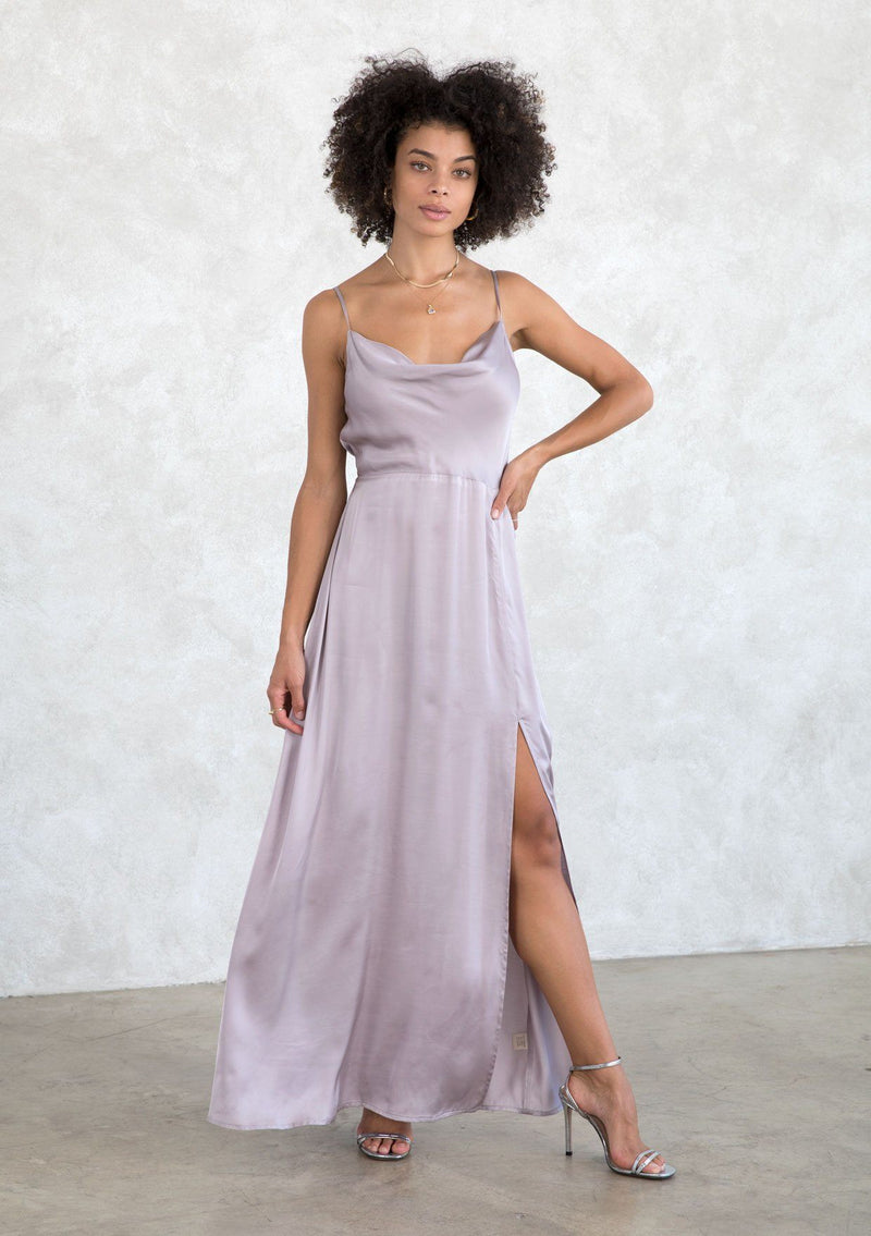 [Color: Lilac Mist] A model wearing a sexy silky lilac purple cowl neck maxi dress. With a half elastic waist at the back, a side slit, and an adjustable strappy back. 