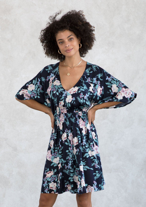 [Color: Navy/Lilac] A model wearing a navy and lilac purple silky floral mini dress designed in a classic bohemian silhouette. With short flutter sleeves, a smocked elastic waist, a tiered paneled skirt, and a v neckline. 