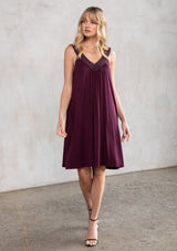 [Color: Fig] A model wearing a purple soft knit, holiday party mini dress with beaded straps, a deep v neckline in front and back, and a flowy swing silhouette. 