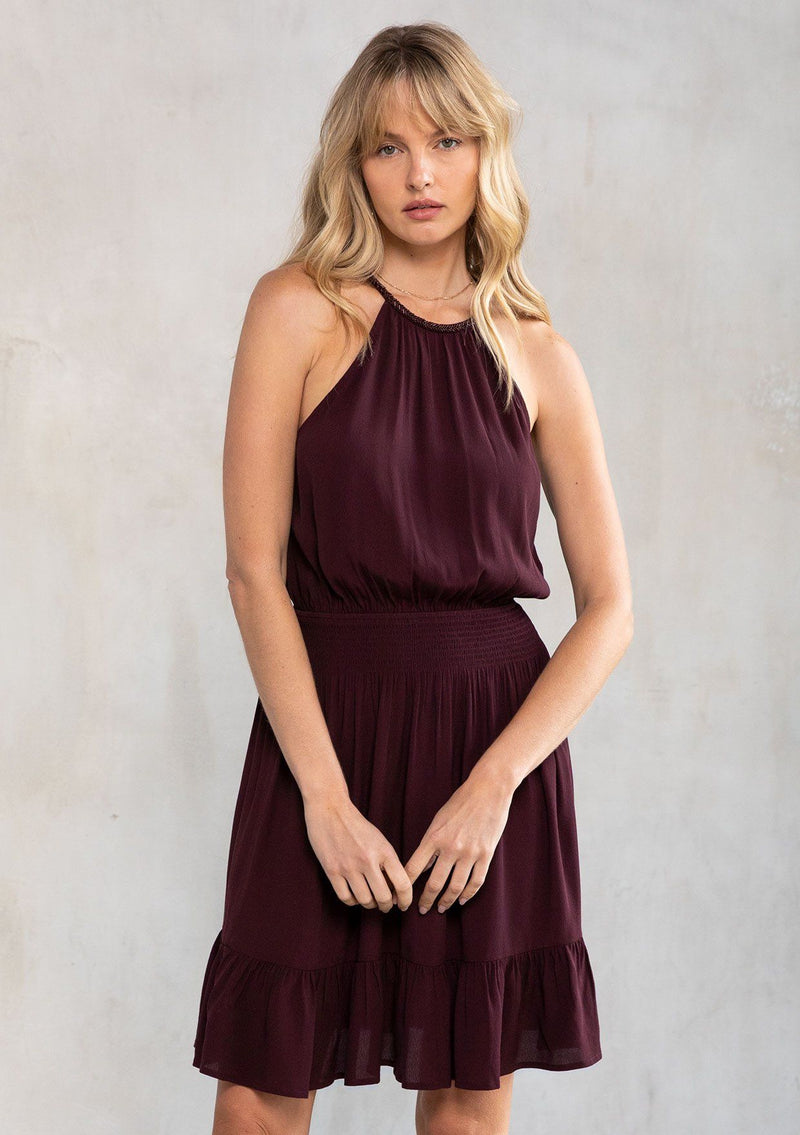[Color: Fig] A model wearing a Holiday ready purple halter mini dress. A sexy dress for your next special occasion, with a beaded accent high neckline, a flattering open back with tassel tie closure, and a smocked elastic waist.