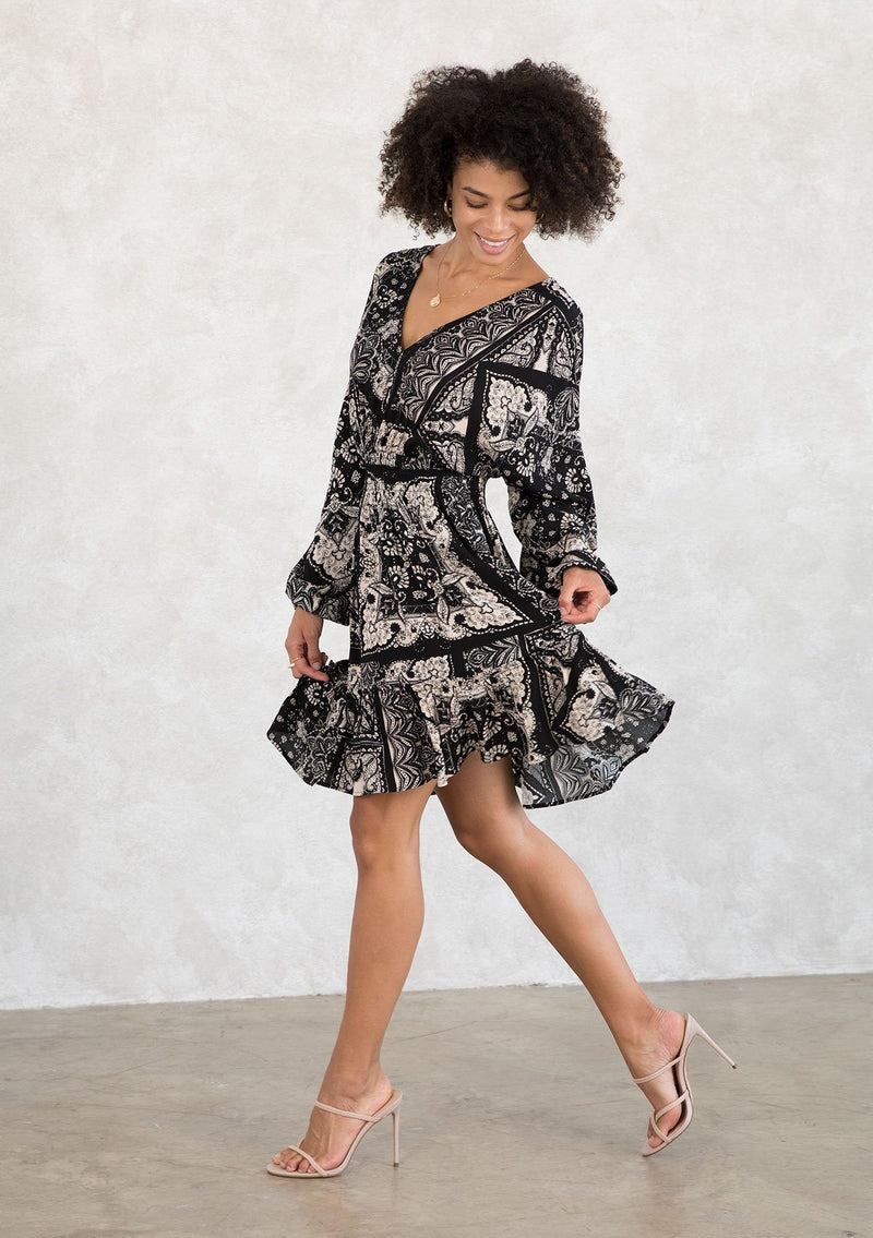 [Color: Black/Taupe] A model wearing a casual bohemian mini dress in a trendy black and taupe bandana print. With voluminous long sleeves, a flattering open back with tassel tie closure, and an elastic waist. 