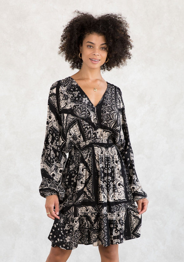 [Color: Black/Taupe] A model wearing a casual bohemian mini dress in a trendy black and taupe bandana print. With voluminous long sleeves, a flattering open back with tassel tie closure, and an elastic waist. 