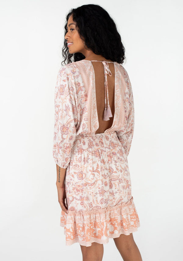 [Color: Natural/Clay] A back facing image of a brunette model wearing a bohemian spring mini dress in a pink floral border print. With voluminous three quarter length sleeves, a v neckline, a smocked elastic waist, a ruffle trimmed flowy tiered skirt, and an open back with tassel tie closure. 