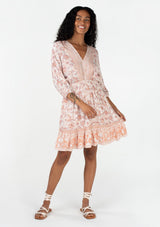 [Color: Natural/Clay] A full body front facing image of a brunette model wearing a bohemian spring mini dress in a pink floral border print. With voluminous three quarter length sleeves, a v neckline, a smocked elastic waist, a ruffle trimmed flowy tiered skirt, and an open back with tassel tie closure. 