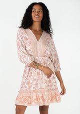 [Color: Natural/Clay] A front facing image of a brunette model wearing a bohemian spring mini dress in a pink floral border print. With voluminous three quarter length sleeves, a v neckline, a smocked elastic waist, a ruffle trimmed flowy tiered skirt, and an open back with tassel tie closure. 