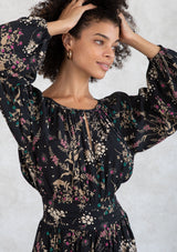 [Color: Black/Jade] A model earing a black and green wildflower print mini dress. With a round neckline and front keyhole detail, an open back with tassel tie closure, a pleated waist detail, and long voluminous sleeves. 