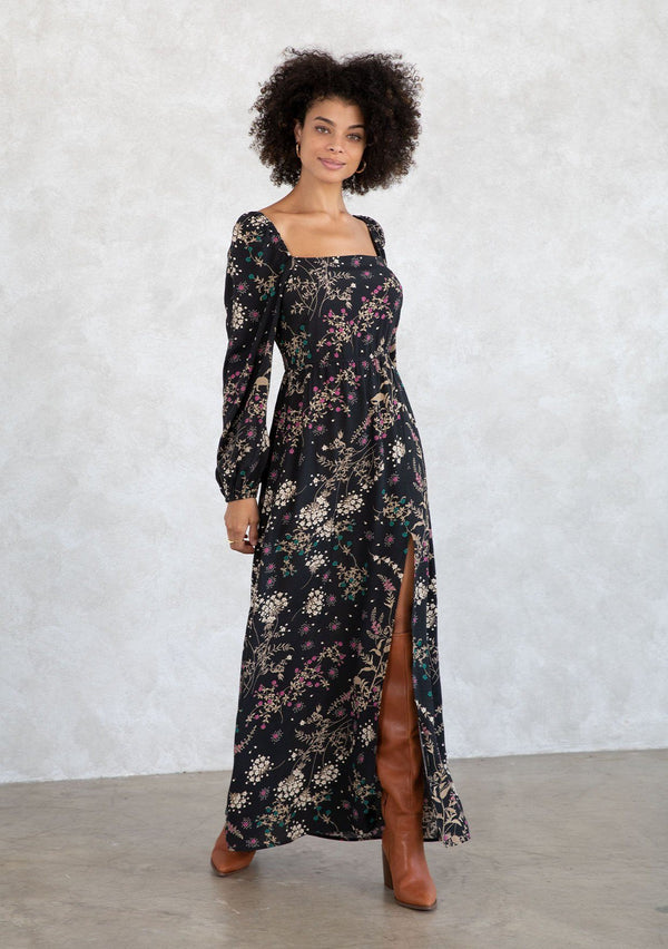 [Color: Black/Jade] A model wearing a bohemian black and green wildflower print maxi dress. With a square neckline, a smocked elastic back, a side slit, and voluminous long sleeves. 