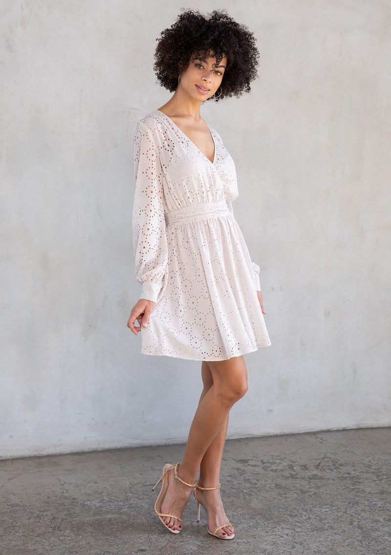 [Color: Bone] A model wearing a bohemian holiday cream colored mini dress in metallic laser cut chiffon. With a surplice v neckline, long sleeves, and a flattering fit and flare silhouette.