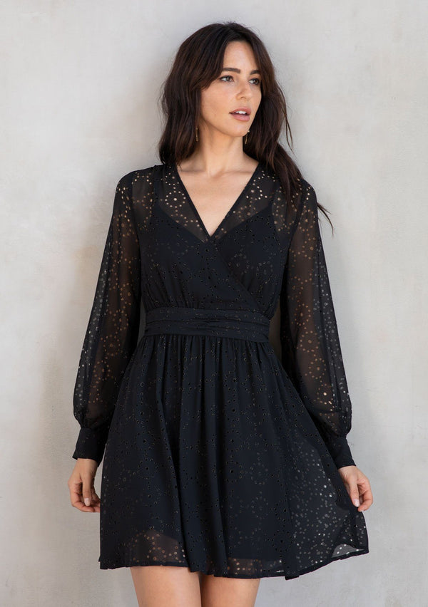 [Color: Black] A model wearing a bohemian holiday mini dress in metallic laser cut chiffon. With a surplice v neckline, long sleeves, and a flattering fit and flare silhouette.