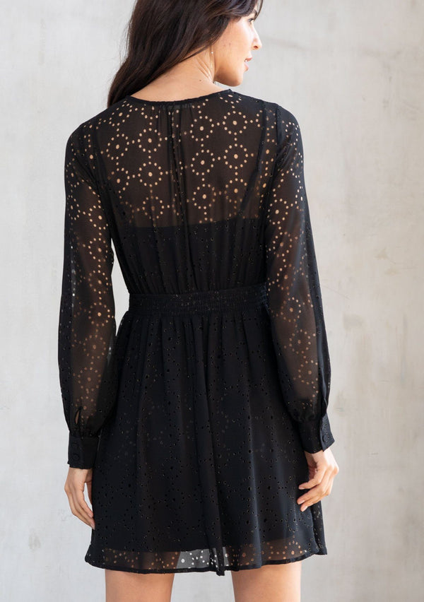 [Color: Black] A model wearing a bohemian holiday mini dress in metallic laser cut chiffon. With a surplice v neckline, long sleeves, and a flattering fit and flare silhouette.