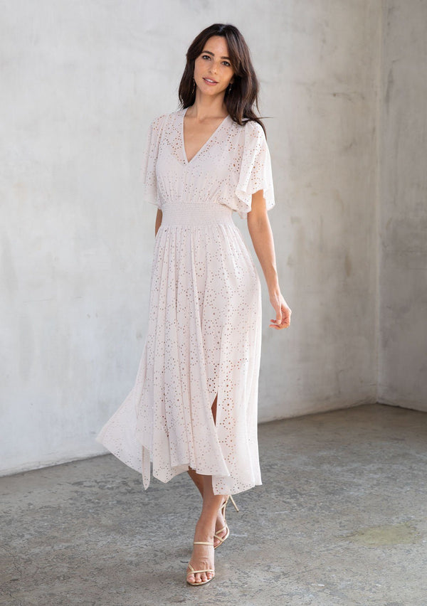 [Color: Bone] A model wearing a dreamy sheer off white chiffon maxi dress with gold trimmed laser cut details throughout. With a flowy skirt, short flutter sleeves, and a shark bite hemline.