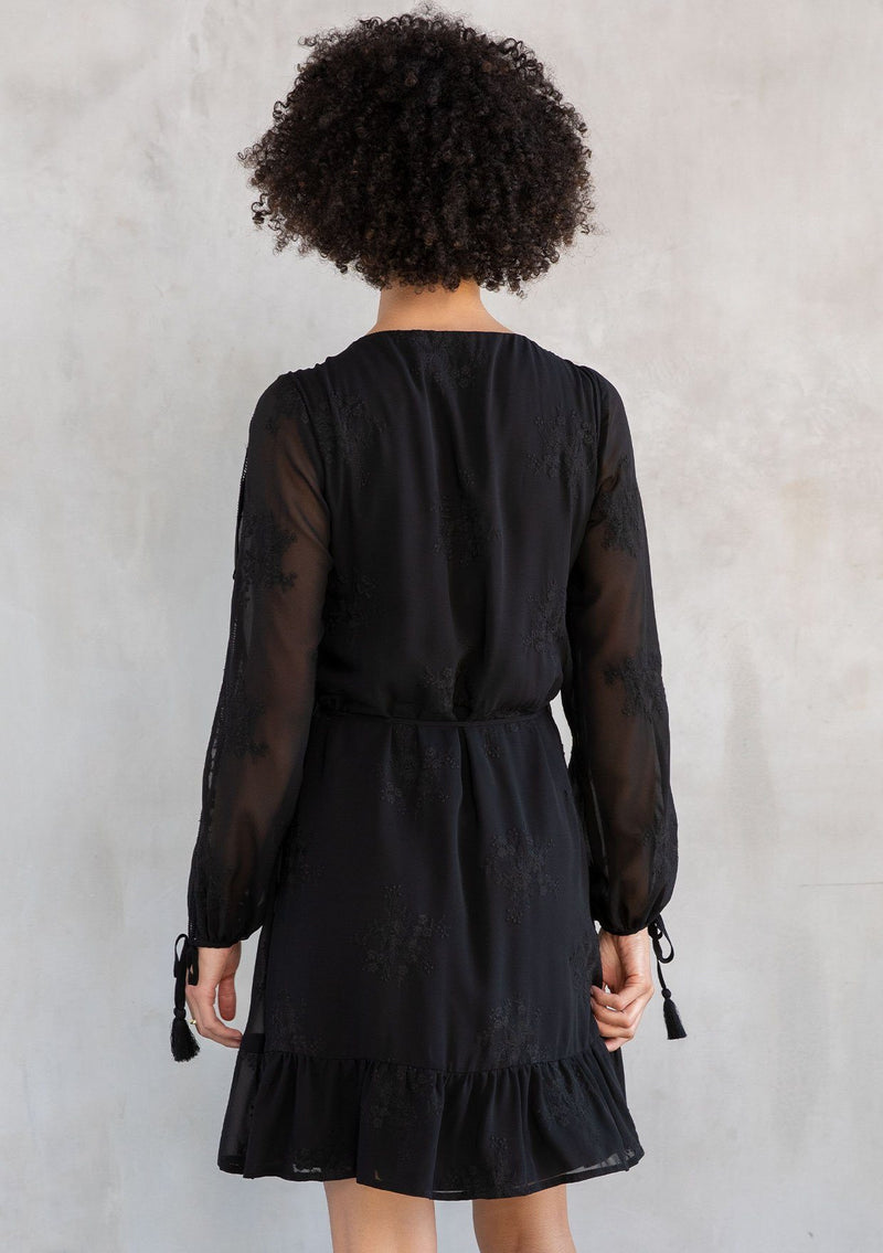 [Color: Black] A model wearing a charming black embroidered chiffon mini wrap dress, with sheer long split sleeves, lattice trim details, and a wrap front with side tie closure.
