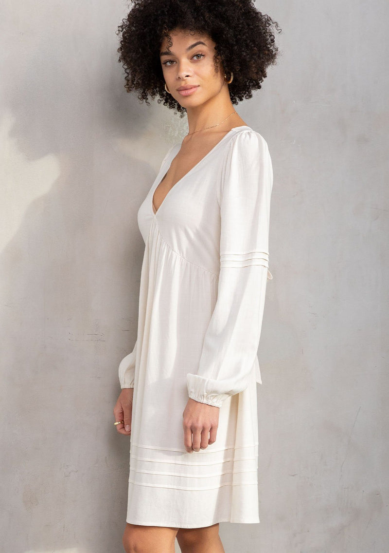 [Color: Vanilla] A model wearing a classic off white bohemian mini dress in a linen blend. With long voluminous sleeves, a tie back detail, and open back detail. 