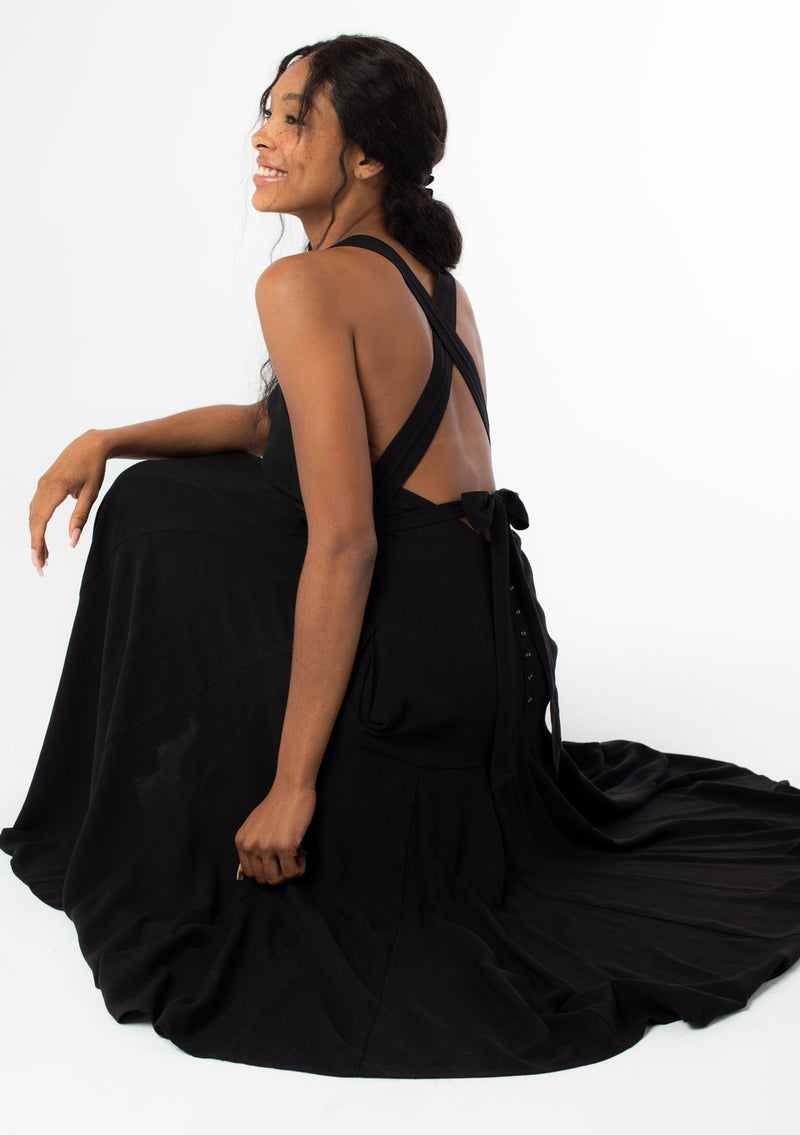 [Color: Black] A sitting side image of a black model wearing a sleeveless black halter maxi dress. A linen blend tank dress with a sexy open back, cross back racer back straps, side pockets, and a back button detail. 