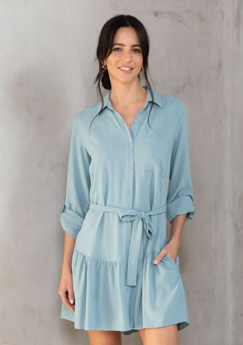 [Color: Dusty Teal] A model wearing a pretty light teal linen blend mini shirtdress. With a flowy tiered skirt, a button front, and long rolled sleeves. 