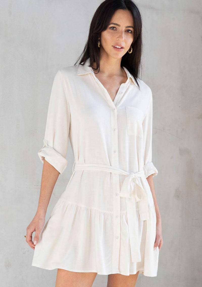 [Color: Vanilla] A model wearing a pretty off white linen blend mini shirtdress. With a flowy tiered skirt, a button front, and long rolled sleeves. 