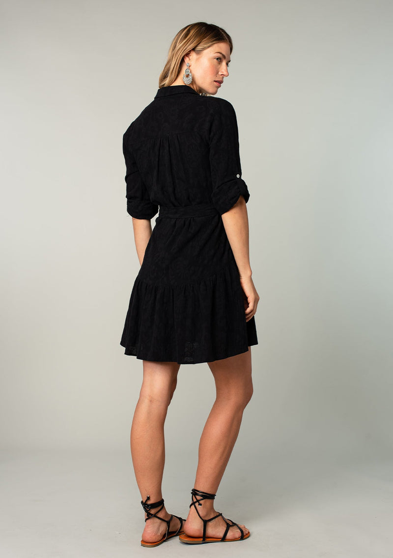 [Color: Black] A back facing image of a blonde model wearing a black cotton jacquard mini shirt dress. An elevated bohemian short dress with a swingy tiered skirt, a button front, a classic collared neckline, a self tie waist belt, and long rolled sleeves with a button tab closure. 