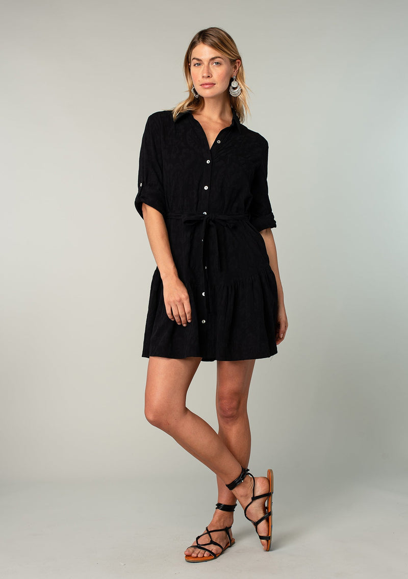 [Color: Black] A full body front facing image of a blonde model wearing a black cotton jacquard mini shirt dress. An elevated bohemian short dress with a swingy tiered skirt, a button front, a classic collared neckline, a self tie waist belt, and long rolled sleeves with a button tab closure. 