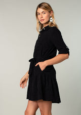 [Color: Black] A side facing image of a blonde model wearing a black cotton jacquard mini shirt dress. An elevated bohemian short dress with a swingy tiered skirt, a button front, a classic collared neckline, a self tie waist belt, and long rolled sleeves with a button tab closure. 