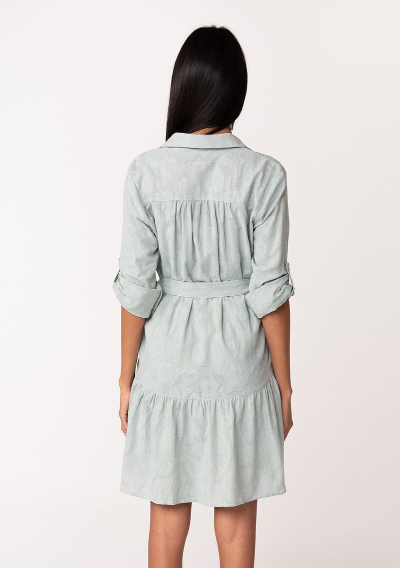 [Color: Sage] A back facing image of a brunette model wearing a light sage green cotton jacquard mini shirt dress. An elevated bohemian short dress with a swingy tiered skirt, a button front, a classic collared neckline, a self tie waist belt, and long rolled sleeves with a button tab closure.