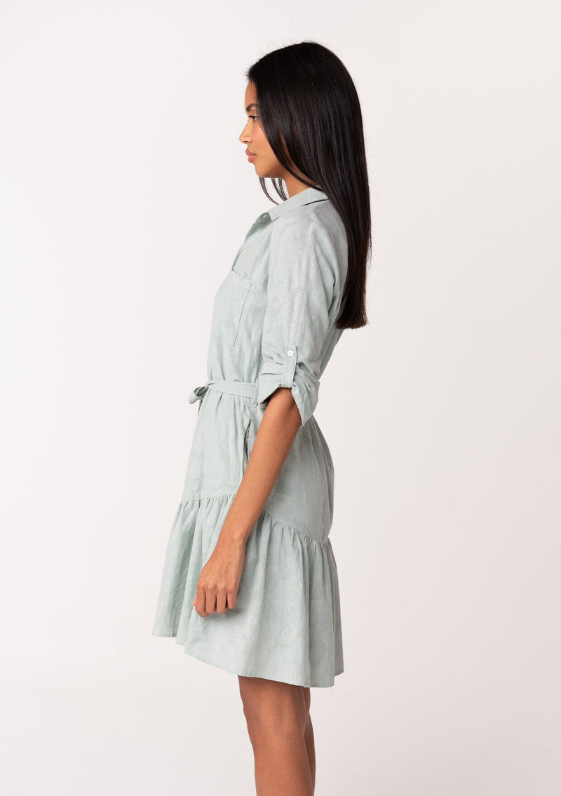 [Color: Sage] A side facing image of a brunette model wearing a light sage green cotton jacquard mini shirt dress. An elevated bohemian short dress with a swingy tiered skirt, a button front, a classic collared neckline, a self tie waist belt, and long rolled sleeves with a button tab closure.