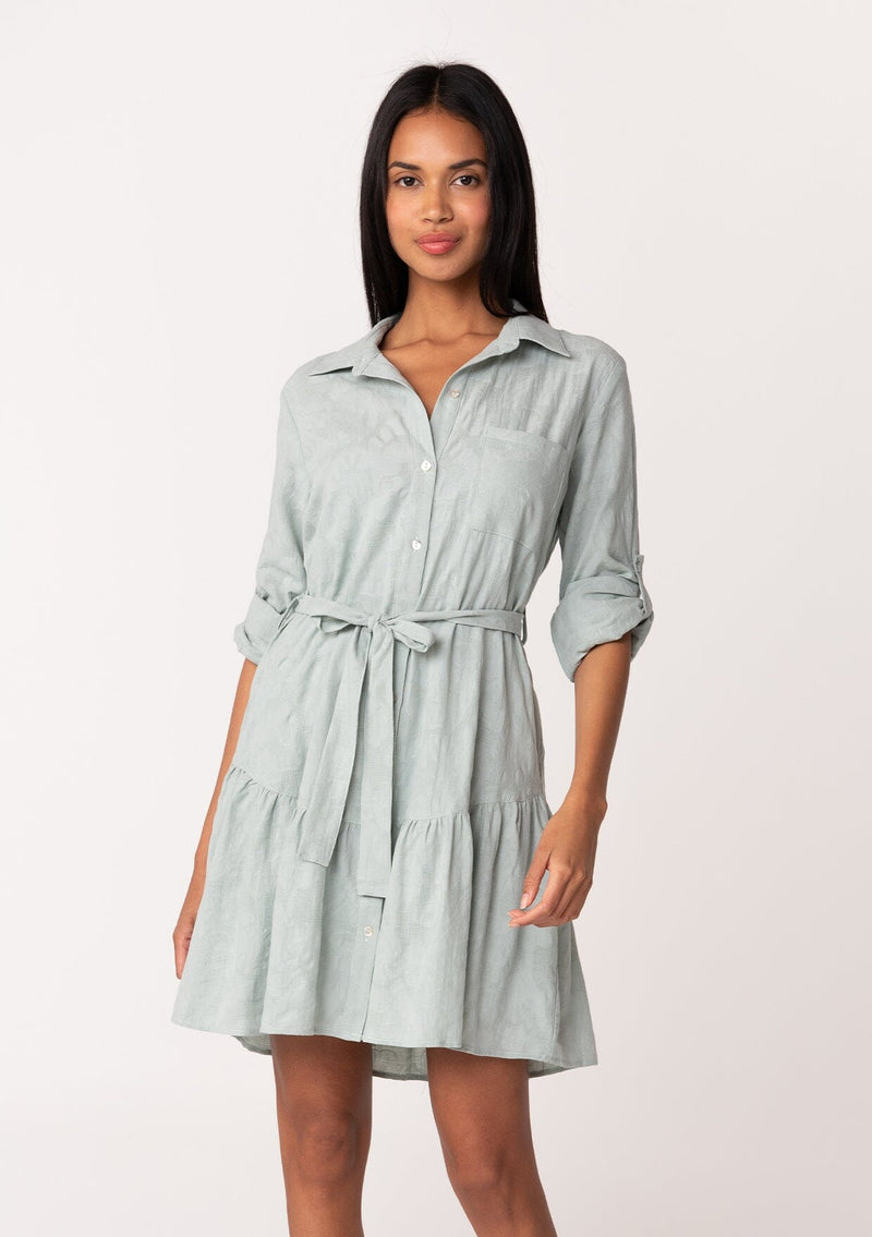 [Color: Sage] A front facing image of a brunette model wearing a light sage green cotton jacquard mini shirt dress. An elevated bohemian short dress with a swingy tiered skirt, a button front, a classic collared neckline, a self tie waist belt, and long rolled sleeves with a button tab closure.