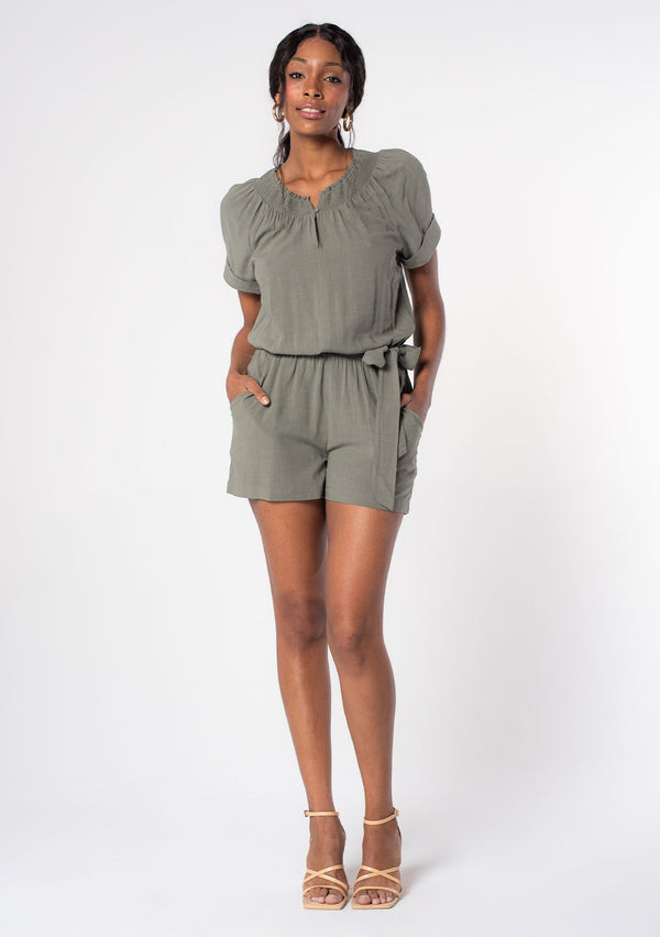 [Color: Olive] A woman wearing an olive green linen blend short romper with short cuffed sleeves, side pockets, and a self tie waist belt.