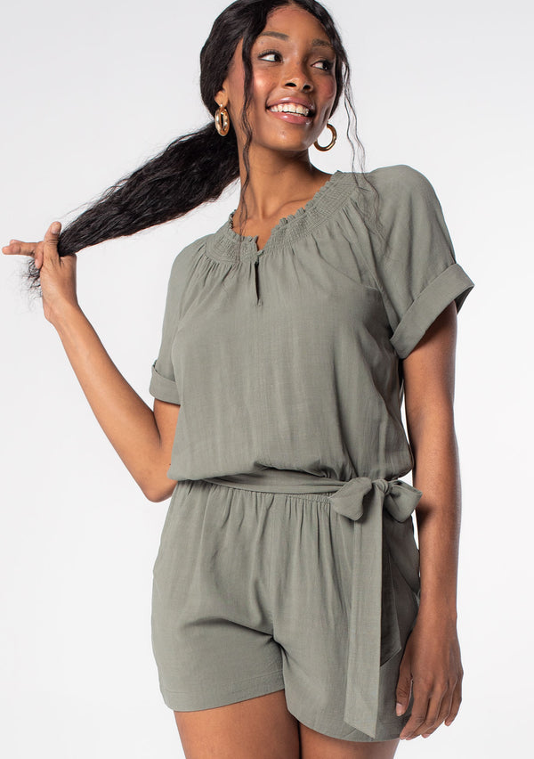 [Color: Olive] A woman wearing an olive green linen blend short romper with short cuffed sleeves, side pockets, and a self tie waist belt.