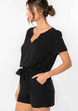 [Color: Black] A woman wearing a black linen blend short romper with short cuffed sleeves, side pockets, and a self tie waist belt. 