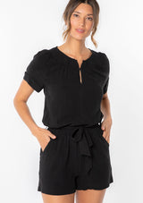 [Color: Black] A woman wearing a black linen blend short romper with short cuffed sleeves, side pockets, and a self tie waist belt. 