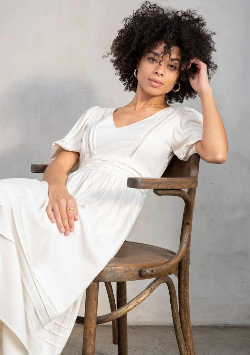 [Color: Vanilla] A model wearing a timeless off white linen blend maxi dress. With short puff sleeves, a flattering pleated waist, and delicate lattice trim. 