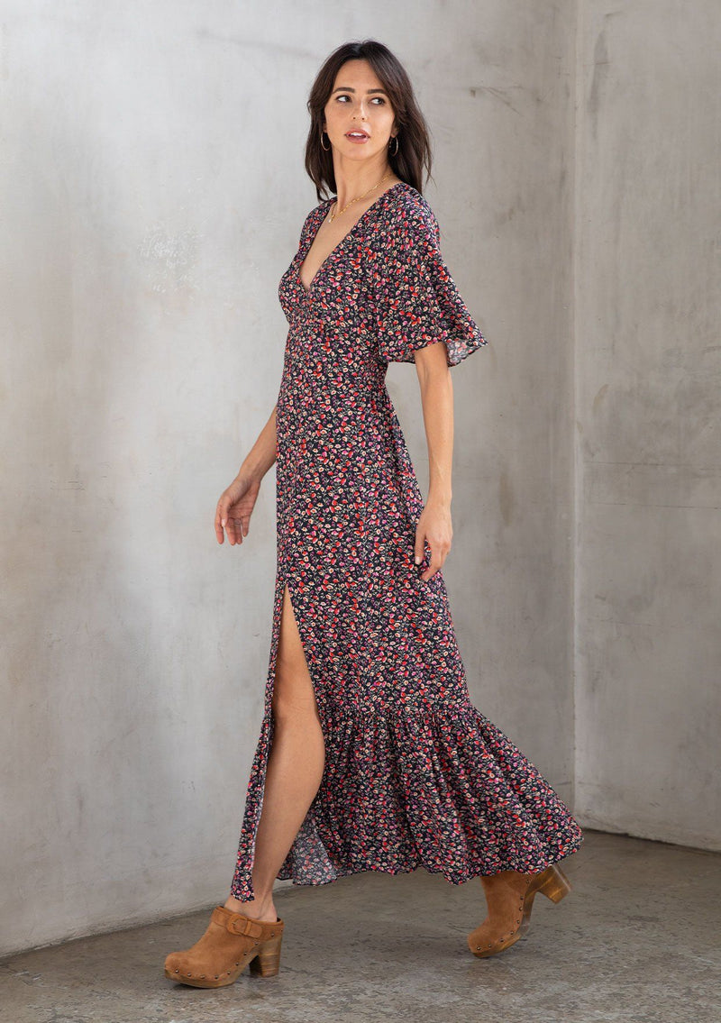 [Color: Black/Fuchsia] A model wearing a timeless flowy bohemian maxi dress in a black and fuchsia ditsy floral print. With short flutter sleeves, a deep v neckline, and a leg baring side slit.