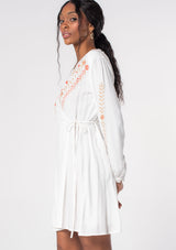 [Color: Ivory/Coral] A model wearing a white mini wrap dress with pink floral embroidery.