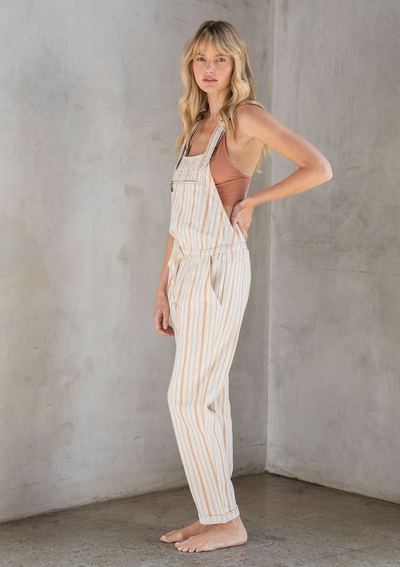 [Color: Natural/Terracotta] A model wearing a classic linen and cotton blend overalls in a terracotta yarn dye stripe. With adjustable straps and a long cuffed leg.