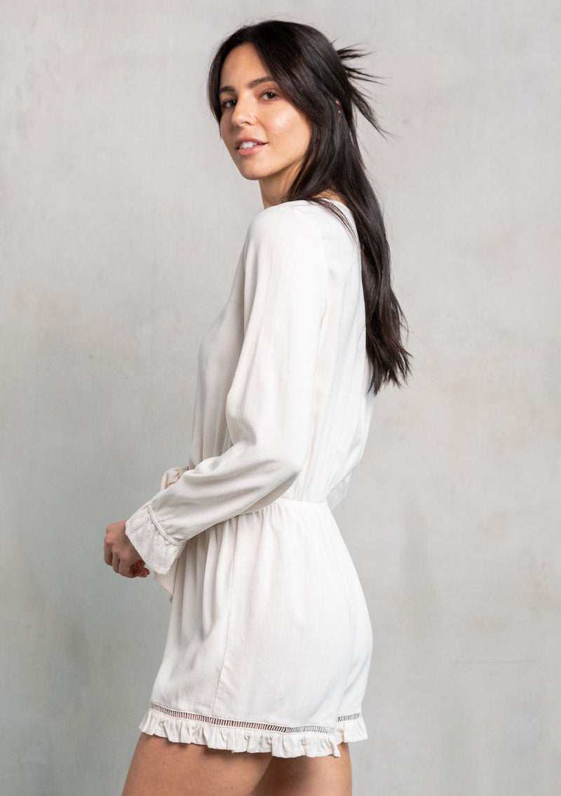 [Color: Vanilla] A model wearing an off white ruffled long sleeve short romper made from a linen blend. With a tie front waist detail. 