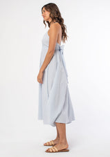 [Color: Periwinkle/White] A woman wearing a cotton blue and white gingham check mid length dress with adjustable spaghetti straps and a tie back detail. 