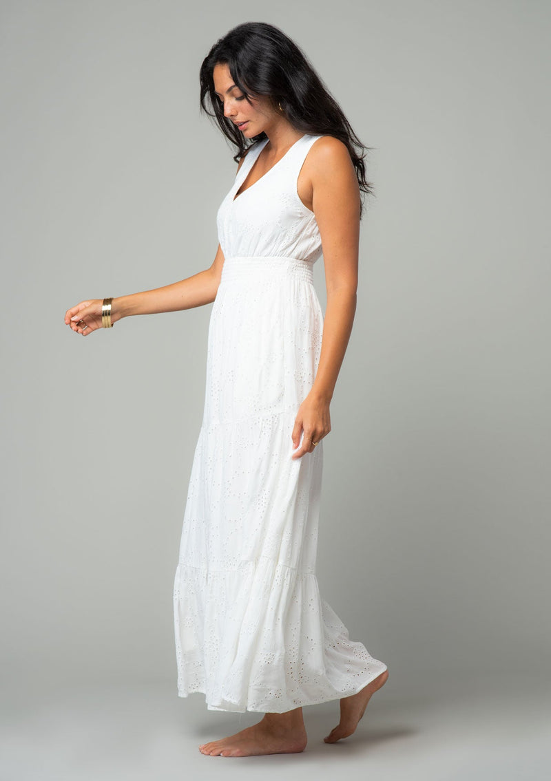 [Color: Off White] A side facing image of a brunette model wearing a sleeveless white bohemian eyelet maxi dress. With a v neckline, a long tiered skirt, and a smocked elastic waist. A classic bohemian white maxi dress for Summer. 