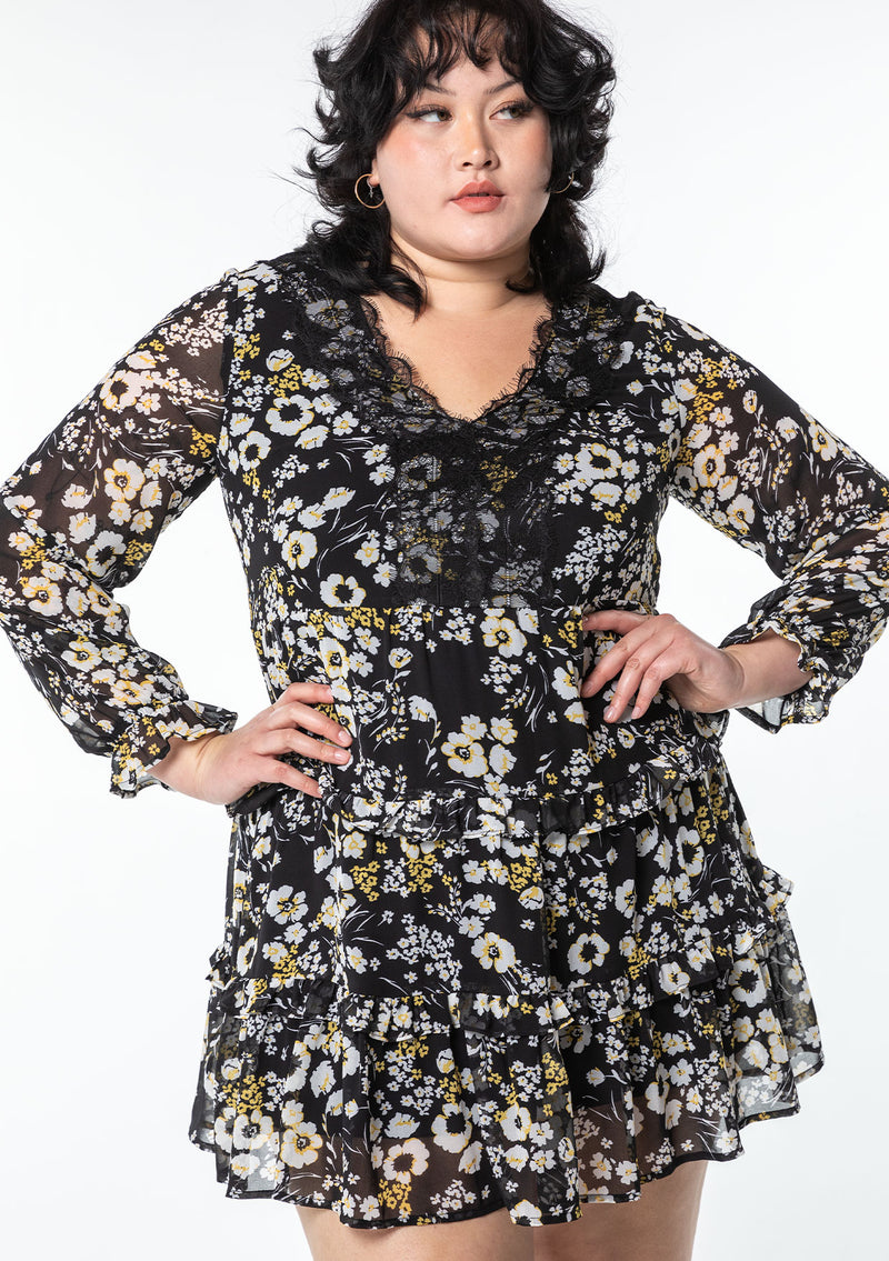 [Color: Black/Lemon] A model wearing an ethereal black chiffon bohemian mini dress with white and yellow floral print. With a lace accent, pretty ruffled trim, and sheer long sleeves.