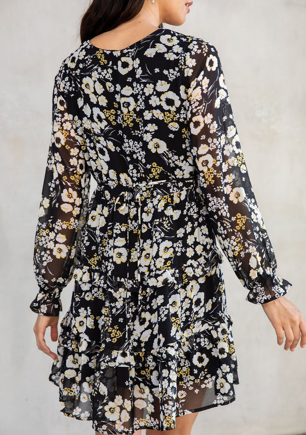 [Color: Black/Lemon] A model wearing an ethereal black chiffon bohemian mini dress with white and yellow floral print. With a lace accent, pretty ruffled trim, and sheer long sleeves. 