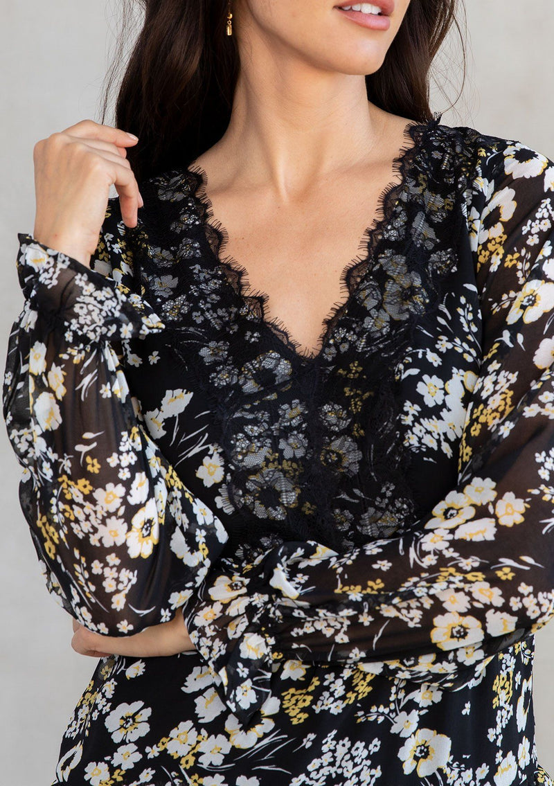 [Color: Black/Lemon] A model wearing an ethereal black chiffon bohemian mini dress with white and yellow floral print. With a lace accent, pretty ruffled trim, and sheer long sleeves. 