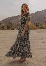 [Color: Black/Lemon] A model wearing a dreamy flowy bohemian maxi dress in a black and yellow floral print. With short puff sleeves, sheer lace accents, and a button front.