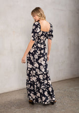 [Color: Black/Natural] A model wearing a stunning black and natural floral maxi dress with short puff sleeves, a sweetheart neckline with adjustable tie front, and a flowy tiered skirt.