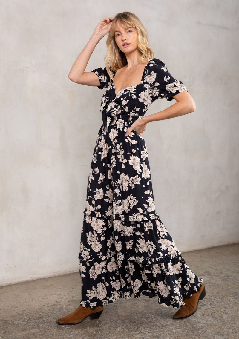 [Color: Black/Natural] A model wearing a stunning black and natural floral maxi dress with short puff sleeves, a sweetheart neckline with adjustable tie front, and a flowy tiered skirt.